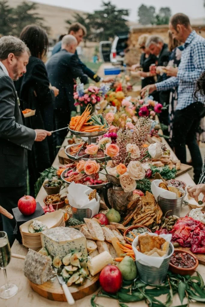 Guests gathered around a long grazing table laden with cheeses, fruits, flowers, and charcuterie outdoors.